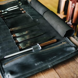 May include: A black leather knife roll with a zipper closure and a brown wooden handle. The roll is filled with various knives and a sharpening steel.