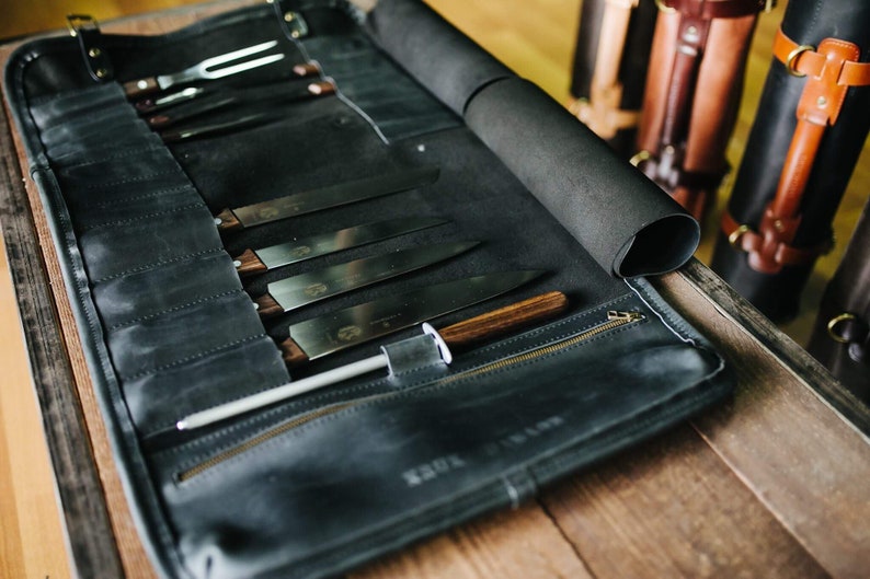 May include: A black leather knife roll with a zipper closure and a brown wooden handle. The roll is filled with various knives and a sharpening steel.