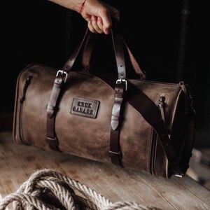 May include: A brown leather duffel bag with a rectangular patch that reads "TRUE GARAGE". The bag has two handles and a shoulder strap, secured with silver buckles. The bag is held up by a person's hand.