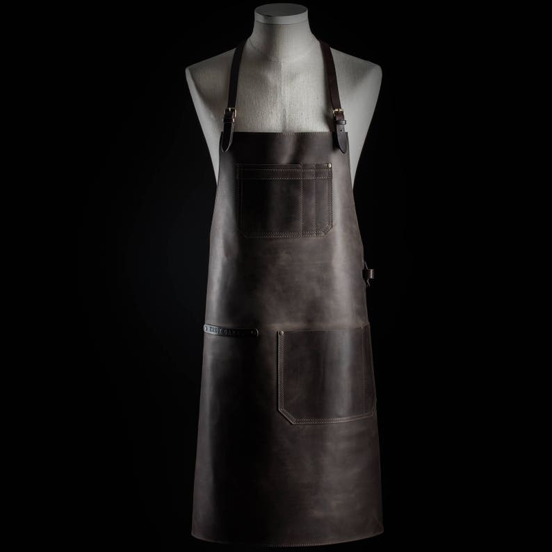 May include: A brown leather apron with adjustable straps and multiple pockets. The apron has a dark brown color and a distressed look.