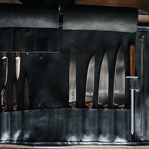 May include: A black leather knife roll with a zippered pocket and the text "KRUK & LARSSON" on the side. The roll is open and contains a variety of knives and a steel.