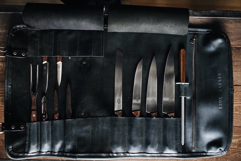 May include: A black leather knife roll with a zippered pocket and the text "KRUK & LARSSON" on the side. The roll is open and contains a variety of knives and a steel.