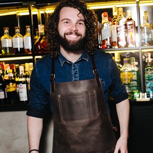 May include: A man with curly brown hair and a beard is wearing a brown leather apron with a large pocket. He is standing in front of a bar with shelves of liquor bottles.