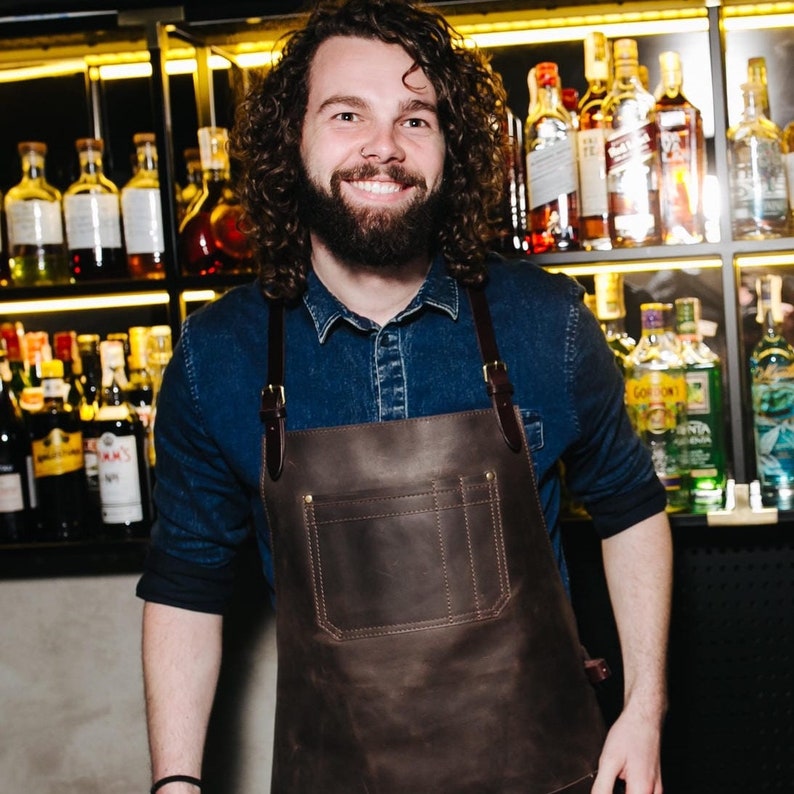 May include: A man with curly brown hair and a beard is wearing a brown leather apron with a large pocket. He is standing in front of a bar with shelves of liquor bottles.