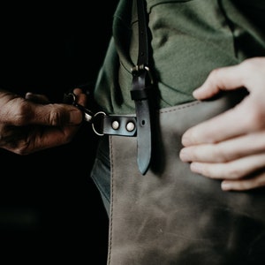 May include: A close-up of a leather apron with a dark brown finish. The apron has a black leather strap with metal hardware. The person is adjusting the strap with their hands. The apron is worn over a green shirt.