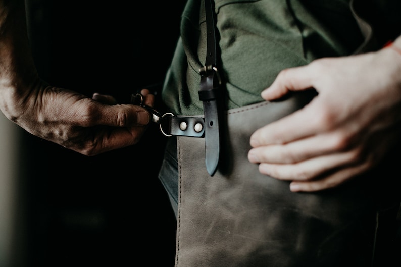 May include: A close-up of a leather apron with a dark brown finish. The apron has a black leather strap with metal hardware. The person is adjusting the strap with their hands. The apron is worn over a green shirt.