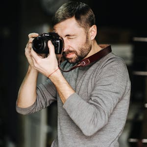 May include: A man wearing a gray sweater is holding a black camera with a brown leather strap and taking a picture.