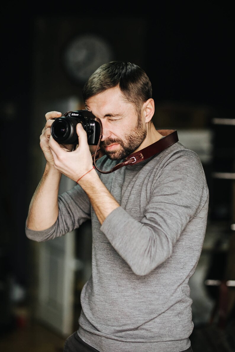 May include: A man wearing a gray sweater is holding a black camera with a brown leather strap and taking a picture.