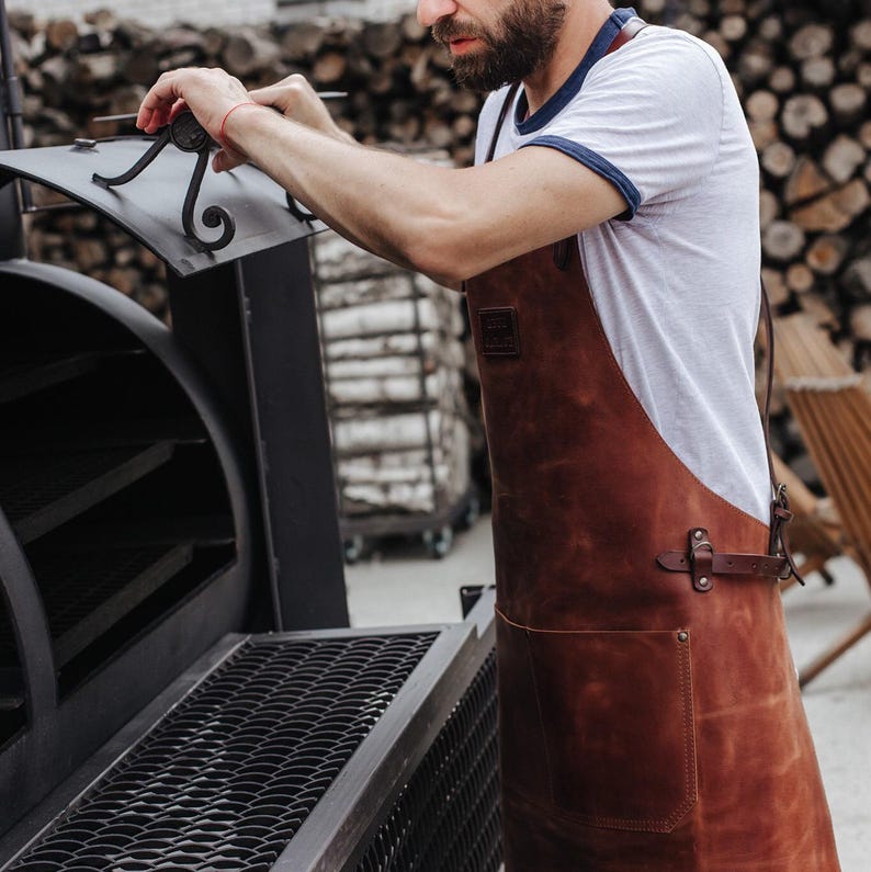 May include: A man wearing a white t-shirt with a blue trim and a brown leather apron is opening the lid of a black metal smoker. The smoker has multiple shelves and a grate on the bottom.