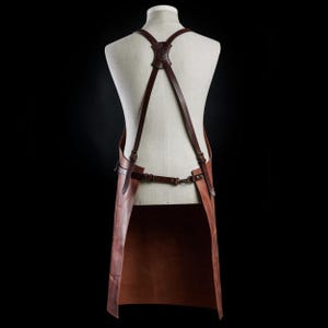 May include: A brown leather apron with adjustable straps and a long bib. The apron is made of thick, durable leather and has a classic design.
