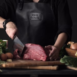 May include: A person wearing a black leather apron with the text "ERUK GARAGE" on it, is cutting a large piece of raw meat with a butcher knife. There are vegetables and other ingredients on the table.