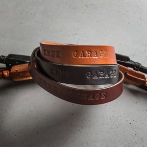 May include: Three leather camera straps in brown, black, and dark brown with the text "KRUK GARAGE" embossed on each strap.