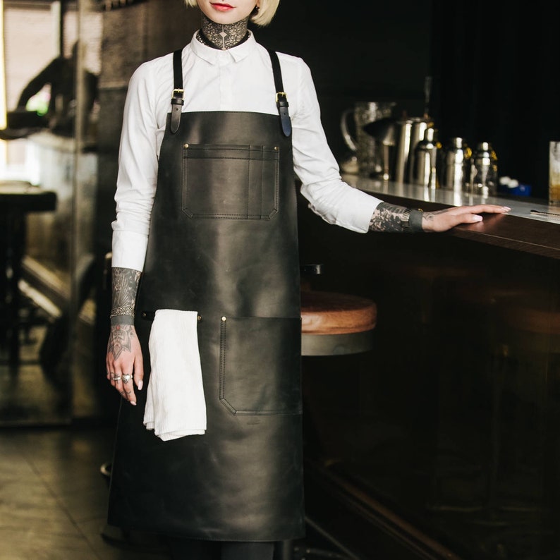 May include: A black leather apron with adjustable straps and a large pocket. The apron is worn by a person with tattoos on their arms.