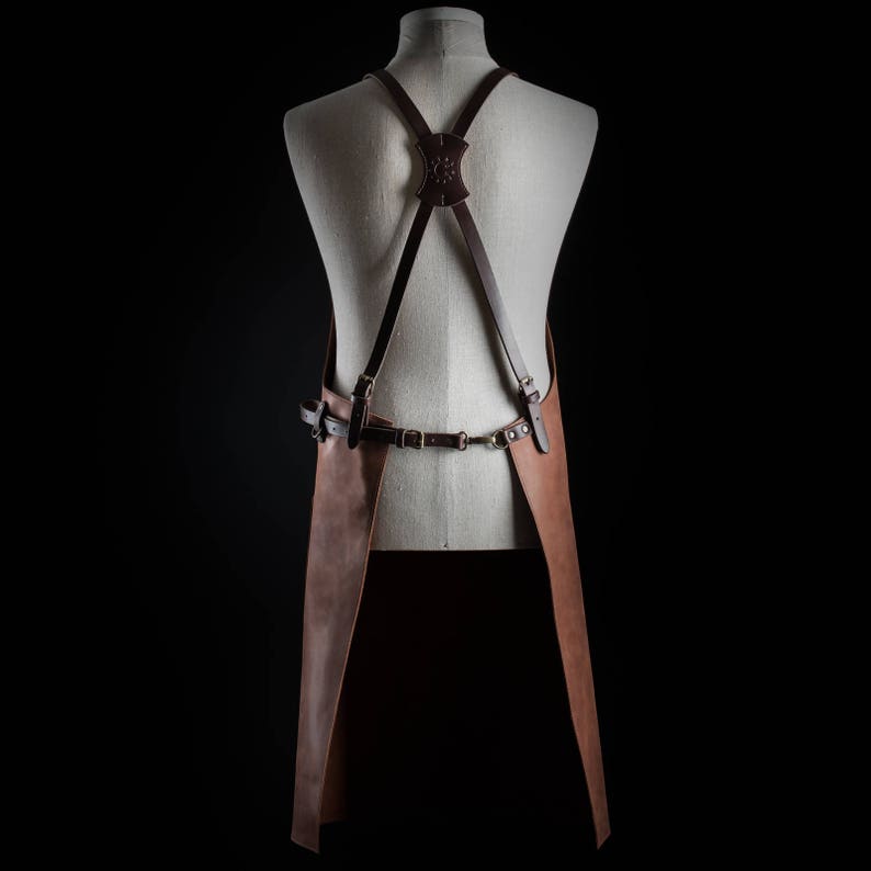 May include: Brown leather apron with adjustable straps and a buckle closure. The apron has a criss-cross strap design in the back.