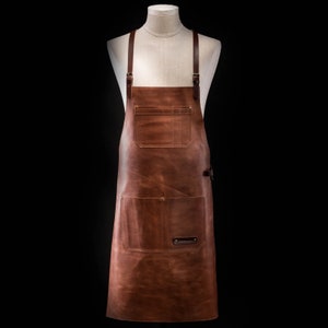 May include: A brown leather apron with two large pockets and adjustable straps. The apron has a small leather patch with the brand name on the front.