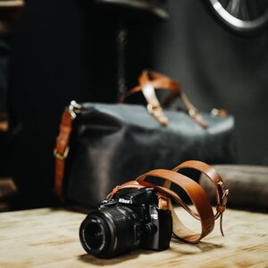 May include: A black camera with a brown leather strap is sitting on a wooden table. The camera has a lens cap on it. There is a brown leather bag in the background.
