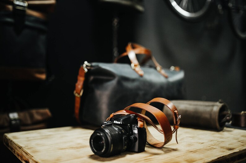 May include: A black camera with a brown leather strap is sitting on a wooden table. The camera has a lens cap on it. There is a brown leather bag in the background.