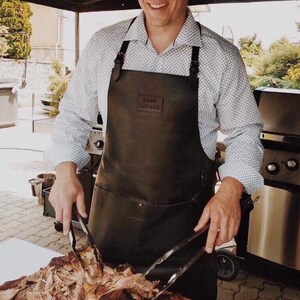 May include: A person wearing a brown leather apron with the text "The Butcher's Apron" on the front, using tongs to lift a large piece of meat.