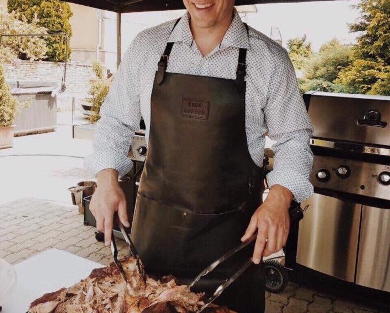 May include: A person wearing a brown leather apron with the text "The Butcher's Apron" on the front, using tongs to lift a large piece of meat.