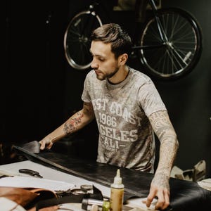 May include: A person working on a leather project in a workshop. They are using a tool to cut a piece of leather. The workshop has a bicycle hanging on the wall and various tools and materials on the work surface.