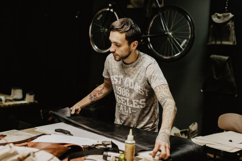 May include: A person working on a leather project in a workshop. They are using a tool to cut a piece of leather. The workshop has a bicycle hanging on the wall and various tools and materials on the work surface.