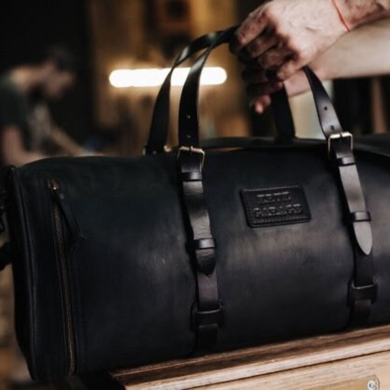 May include: A black leather duffel bag with a top handle and adjustable shoulder straps. The bag has a zippered closure and a leather patch with the text "The  Factory" on the front.