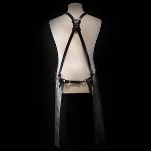 May include: Black leather apron with adjustable straps and a decorative skull accent on the back. The apron features a waist belt with metal hardware and extends down to the knees. The apron is displayed on a mannequin against a black background.