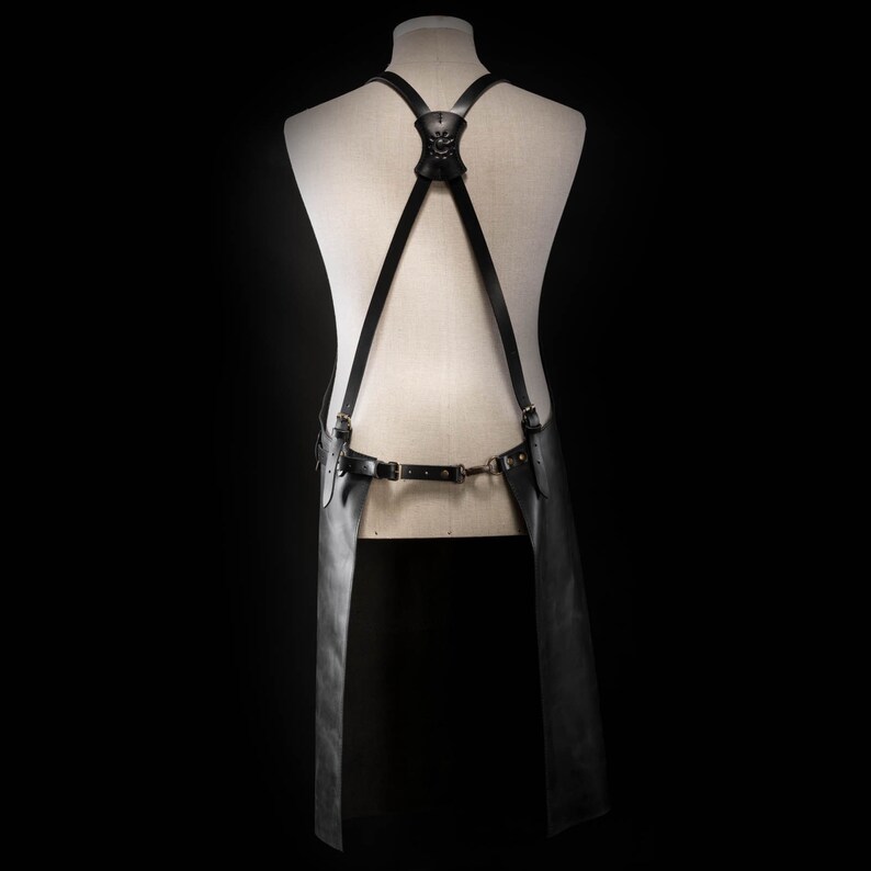 May include: Black leather apron with adjustable straps and a decorative skull accent on the back. The apron features a waist belt with metal hardware and extends down to the knees. The apron is displayed on a mannequin against a black background.