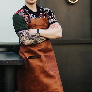 May include: A brown leather apron with a large pocket and a strap across the chest. The apron is worn by a person with a colorful shirt and tattoos on their arms.