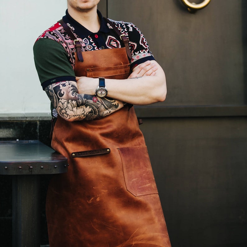 May include: A brown leather apron with a large pocket and a strap across the chest. The apron is worn by a person with a colorful shirt and tattoos on their arms.