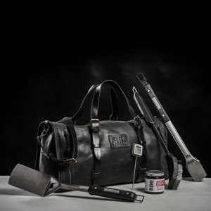 May include: A black leather duffel bag with the text "Crux Garage" on the front. The bag is surrounded by grilling tools, including a spatula, tongs, a grill brush, and a meat thermometer.