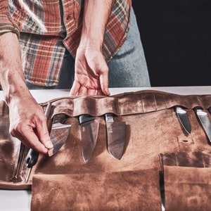 May include: A brown leather roll with multiple pockets for holding knives and other tools. The roll is unrolled and the knives are visible.