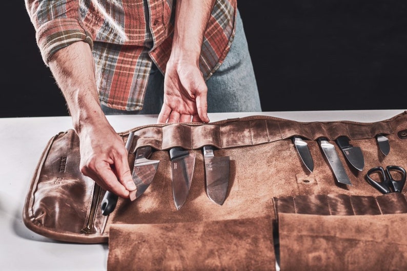 May include: A brown leather roll with multiple pockets for holding knives and other tools. The roll is unrolled and the knives are visible.