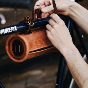 May include: A brown leather bicycle handlebar bag with a zipper closure and the word "KRUX" embossed on the front. The bag is attached to a blue bicycle handlebar with a leather strap.