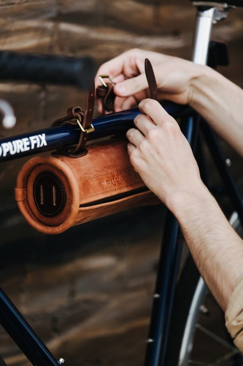 May include: A brown leather bicycle handlebar bag with a zipper closure and the word "KRUX" embossed on the front. The bag is attached to a blue bicycle handlebar with a leather strap.