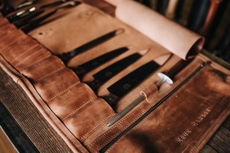 May include: A brown leather knife roll with a zipper closure and the text "KRUIK GARAGE" embossed on the front. The roll is open and contains several knives.