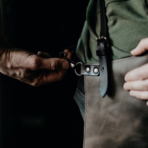 May include: A person wearing a green shirt and a brown leather apron with black straps and silver buckles. The apron has a large pocket on the front.