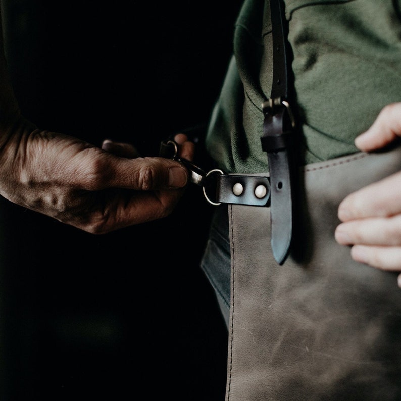 May include: A person wearing a green shirt and a brown leather apron with black straps and silver buckles. The apron has a large pocket on the front.