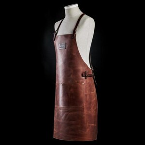 May include: A brown leather apron with an adjustable strap and a large pocket. The apron has a logo that says "The  Barnyard".