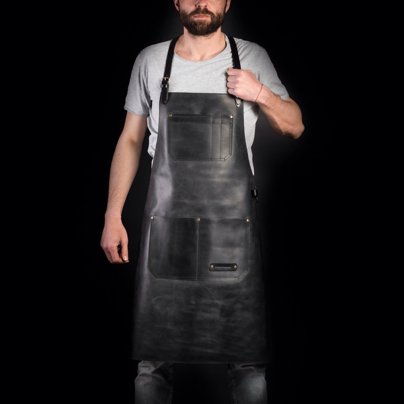 May include: A full-length, dark gray leather apron with two large front pockets and a chest pocket. The apron has adjustable straps and brass-colored rivets. The leather has a distressed appearance. The apron is designed for use in a kitchen or workshop.