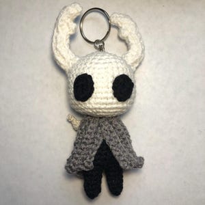 May include: White crocheted keychain with black eyes and a gray and black cloak. The keychain is shaped like a character from the video game Hollow Knight.