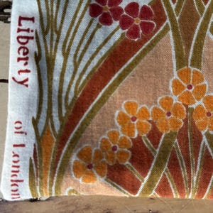 May include: A piece of fabric featuring a floral pattern with red and orange flowers on a cream background. The text Liberty of London is printed in red along the left edge. The design includes stylised green stems and geometric shapes.