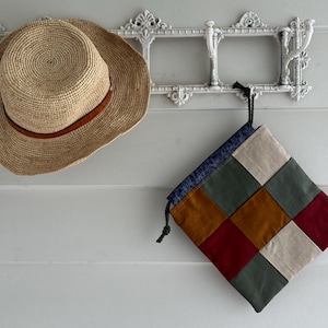 Puede incluir: Un sombrero de paja tejido con una banda de cuero marrón cuelga de un gancho decorativo blanco en la pared. Una bolsa de retazos con cuadrados de tela verde, beige, roja y dorada está suspendida debajo. La bolsa tiene una parte superior azul estampada y un cordón negro.