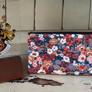 May include: A rectangular fabric pouch featuring a dense floral pattern in shades of red, blue, white, and brown with a black zip closure. Beside it stands a clear acrylic plaque displaying the text Fabrics by Liberty of London.