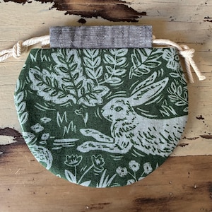 May include: A round drawstring pouch featuring a dark green and light green print of a rabbit surrounded by foliage and flowers. The pouch has a dark grey fabric top band and cream-coloured twisted rope drawstrings on both sides for closure.