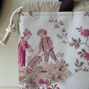 Könnte beinhalten: Tragetasche mit Vintage-Print, der zwei Kinder mit Schubkarre und Blumenelementen in Rosa, Beige und Braun zeigt. Die Tasche hat einen braunen Griff und einen Kordelzugverschluss.