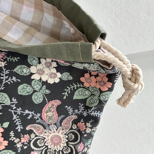 May include: A drawstring bag featuring a dark grey exterior with a multi-coloured floral and paisley print in shades of pink, green, and white. The bag has an olive green top border and a beige and white gingham interior lining with a thick rope cord.