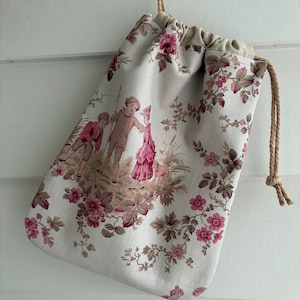 May include: A drawstring pouch made of light grey fabric featuring a repeating toile pattern in shades of pink and brown. The design depicts two children and a small dog in a garden setting, surrounded by floral motifs and vines with a beige cord.