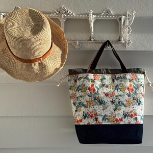 May include: A straw hat with a brown leather band and a tote bag featuring a white fabric with a colourful floral and botanical print, a solid dark blue bottom panel, dark brown handles, and white drawstring cords on the sides.