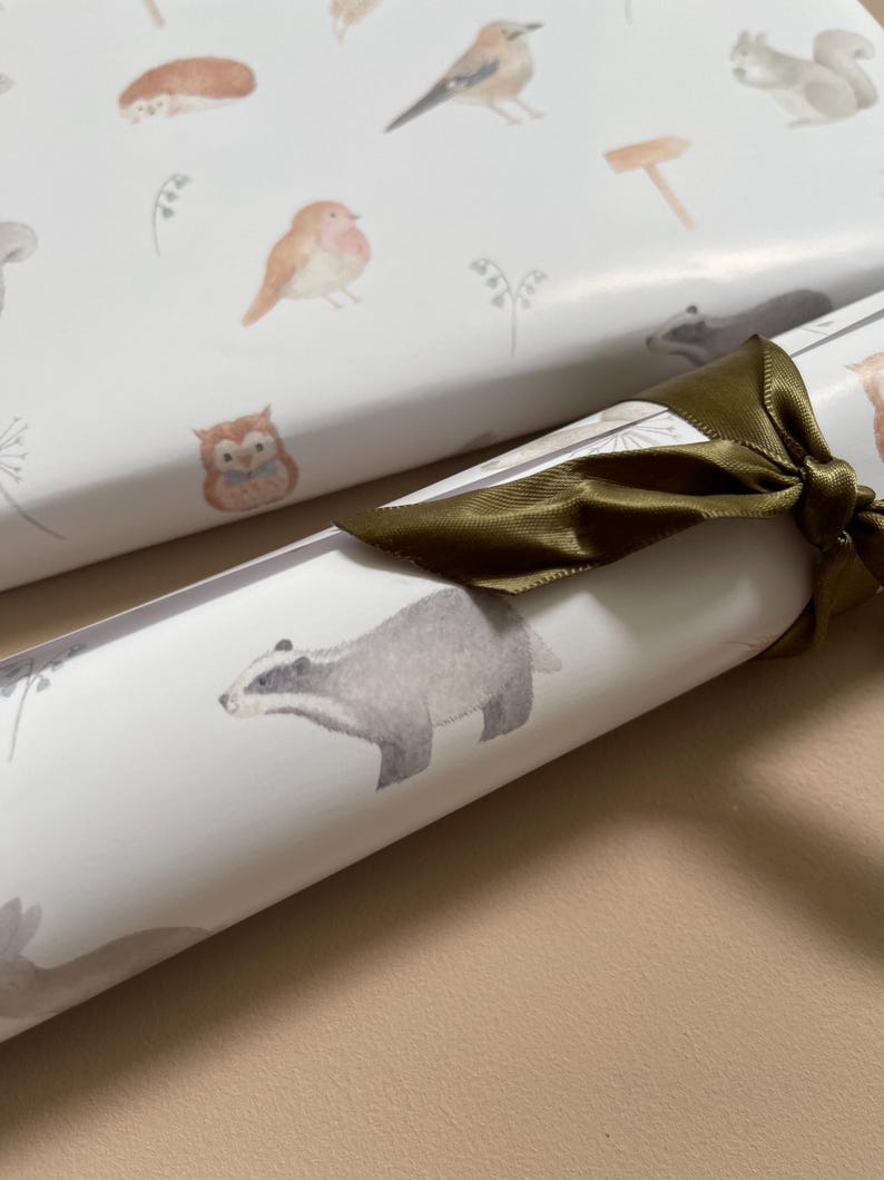 May include: A roll of white wrapping paper with a woodland animal theme. The paper features illustrations of a badger, a squirrel, a bird, an owl, and a robin. The paper is tied with a green ribbon.
